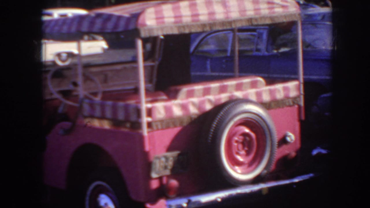 1961 Hawaii home-movie frame showing a pink canopy jeep with a rear spare tire in a sunny parking lot.