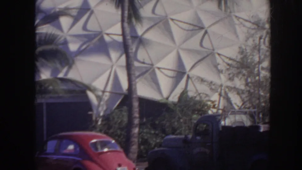 1961 Hawaii frame with palm trees, parked vehicles, shrubs, and a large geometric dome structure in the background.