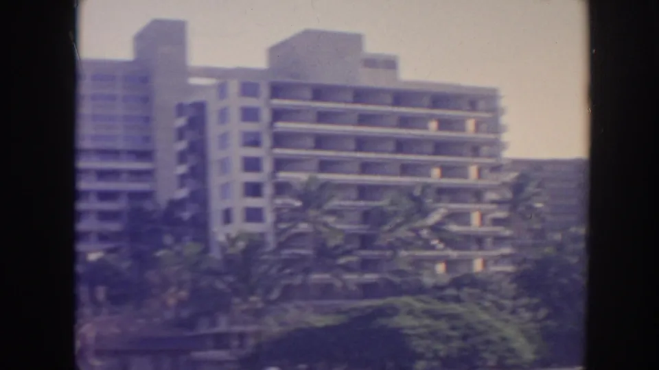 1961 Hawaii frame with a balconied modern building rising behind palm trees and lush landscaping.