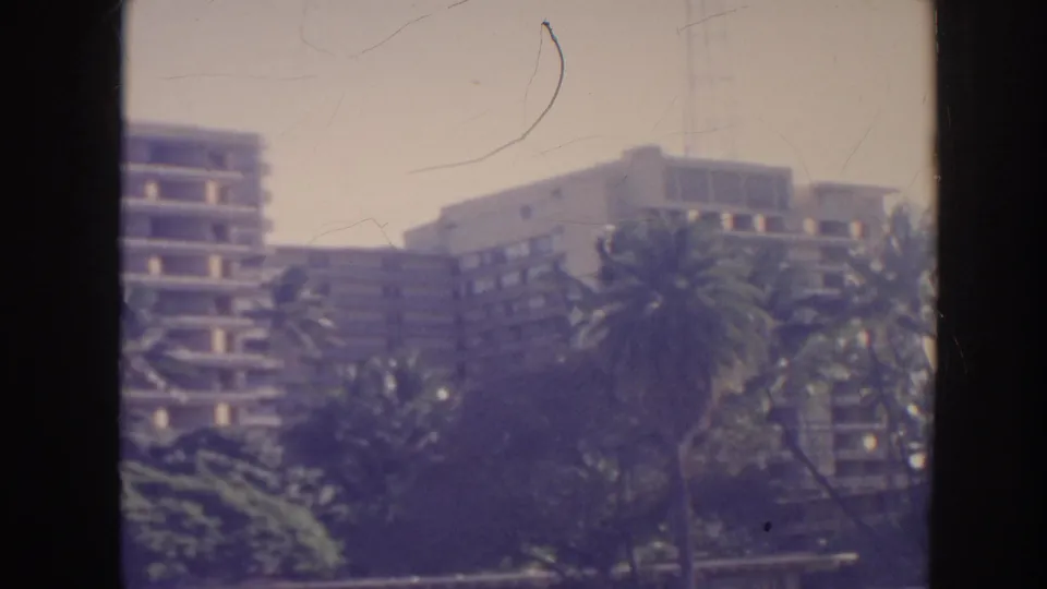 1961 Hawaii 8mm view of palm trees and shrubs partially blocking a multi-story building.