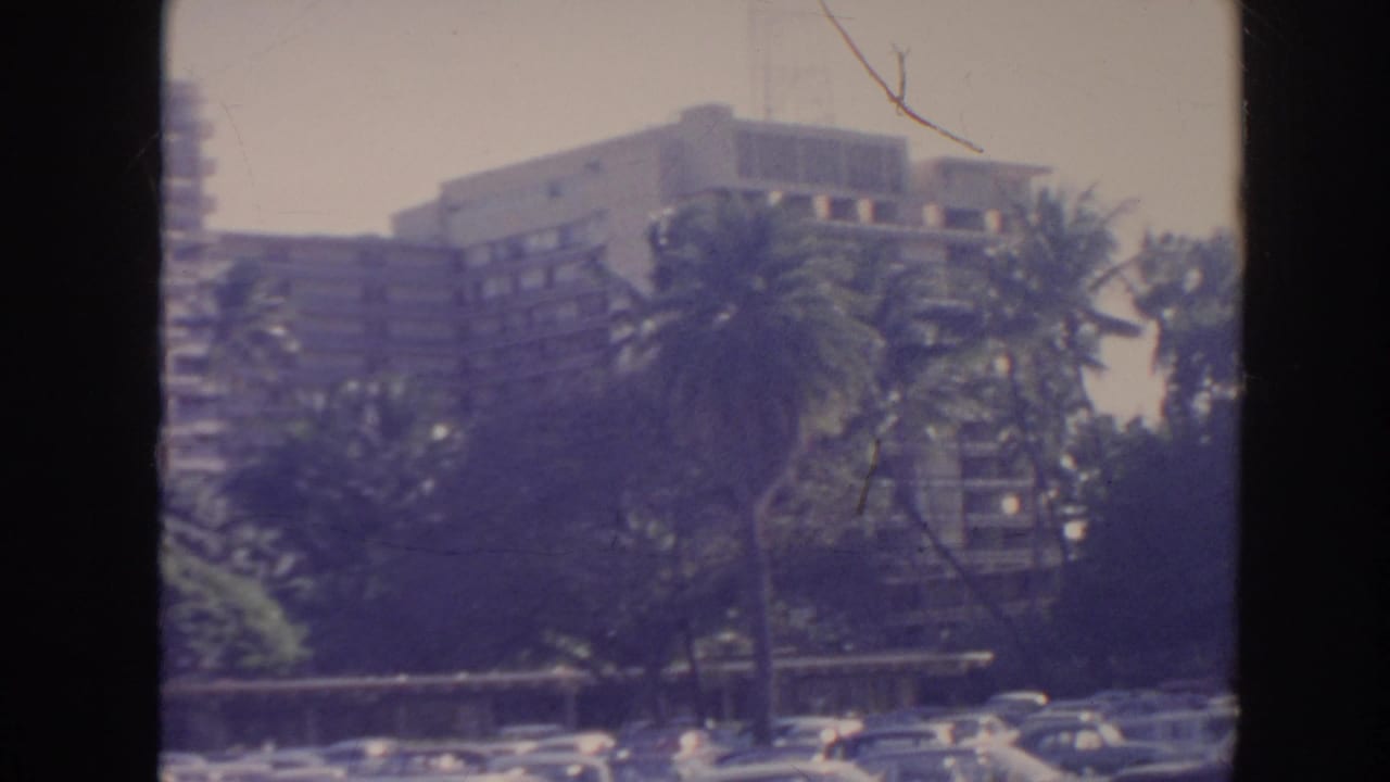 1961 Hawaii home-movie frame showing a resort-like building framed by palms, greenery, and parked cars under a bright sky.