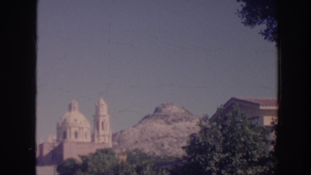 1961 Hawaii home-movie frame with twin church domes, bell towers, trees, and a rocky mountain ridge in the background.