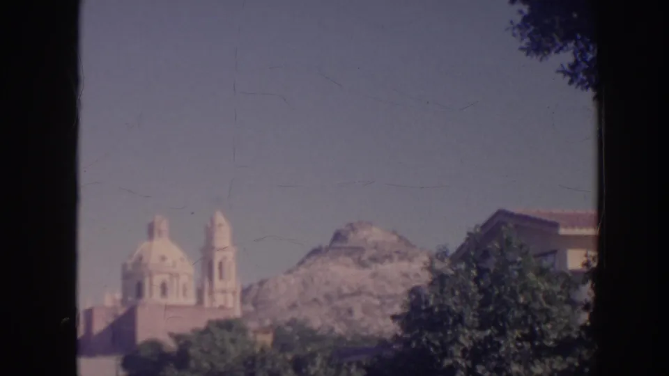 1961 Hawaii home-movie frame with twin church domes, bell towers, trees, and a rocky mountain ridge in the background.