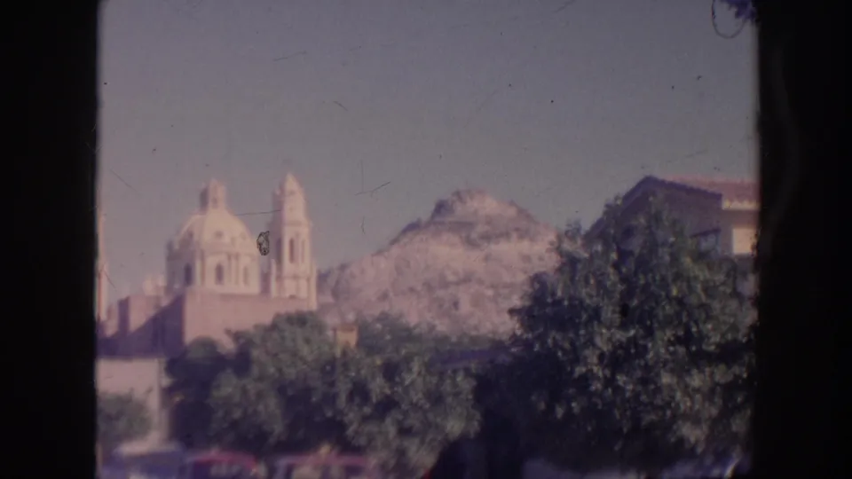 1961 Hawaii 8mm frame showing the church below a mountain ridge, with dense trees and a soft red traffic blur near the bottom edge.