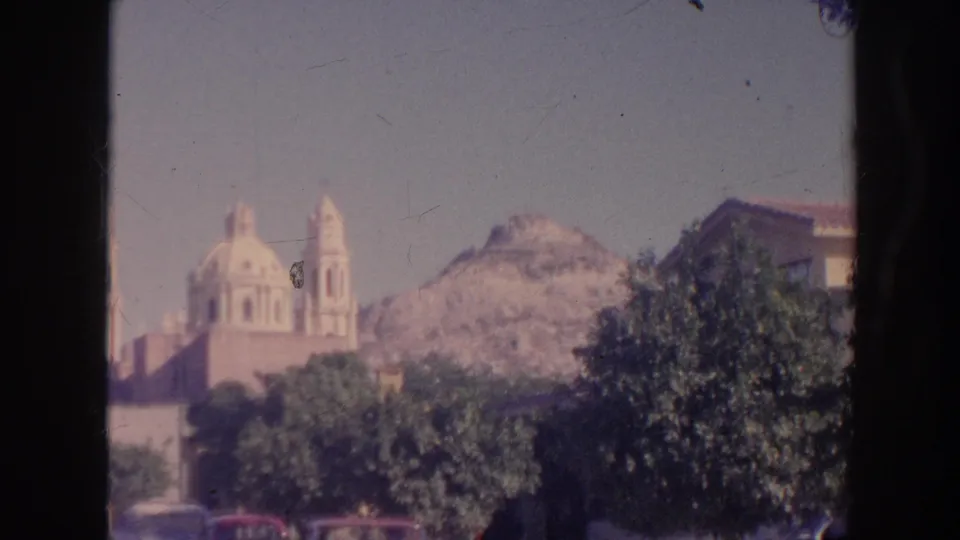 1961 Hawaii home-movie frame of a distant domed church with bell tower, dense trees, pale sky, and rocky mountain backdrop.