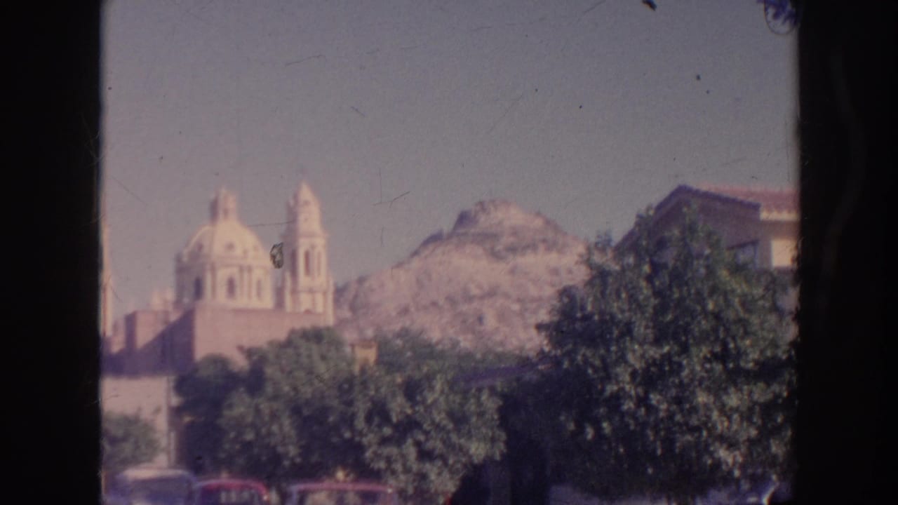 1961 travel footage frame showing a domed church or temple-like exterior, rocky hillside, leafy trees, and parked cars.