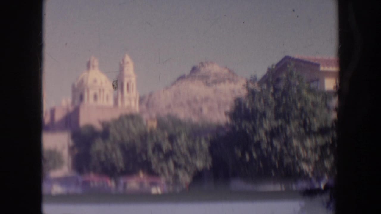 Soft 8mm frame of a domed church or temple-like building near a rocky hill, partly obscured by trees and distance.