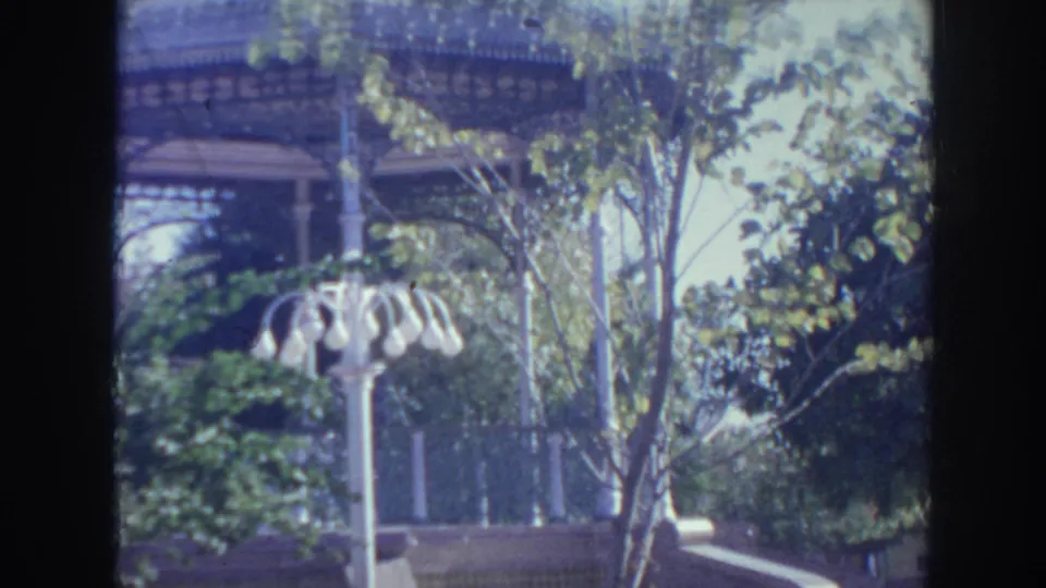 1961 Hawaii 8mm frame showing an ornate gazebo through trees, with shrubs, a low wall, and a white multi-globe lamppost in the foreground.
