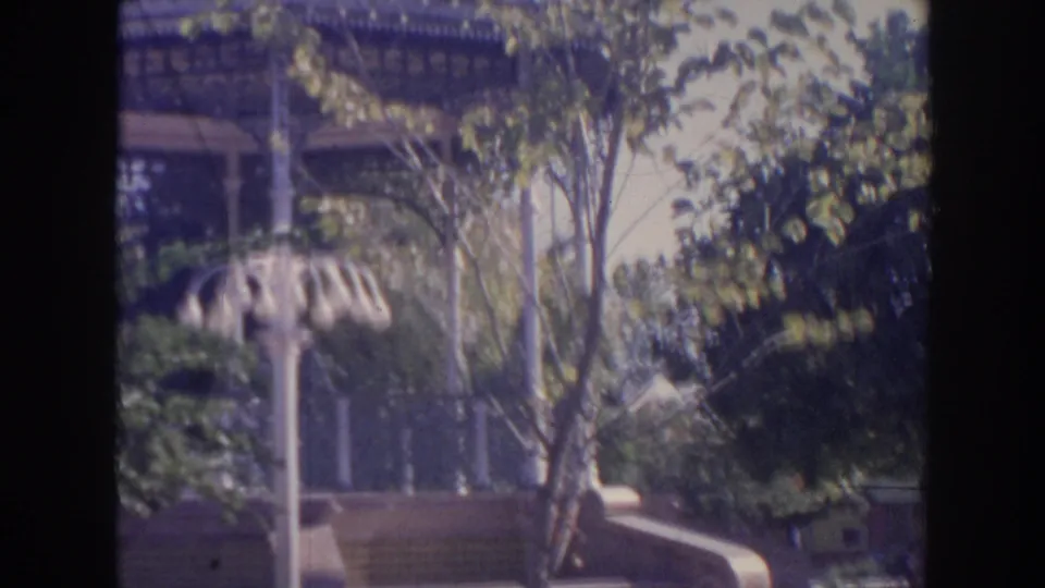 1961 Hawaii home-movie frame with a shaded gazebo partly obscured by trees, dense planting, a curved wall, and a white multi-globe lamppost.