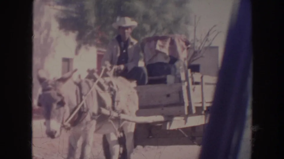 1961 8mm frame showing a man in a light hat seated on a donkey-drawn cart carrying goods along a dusty road.