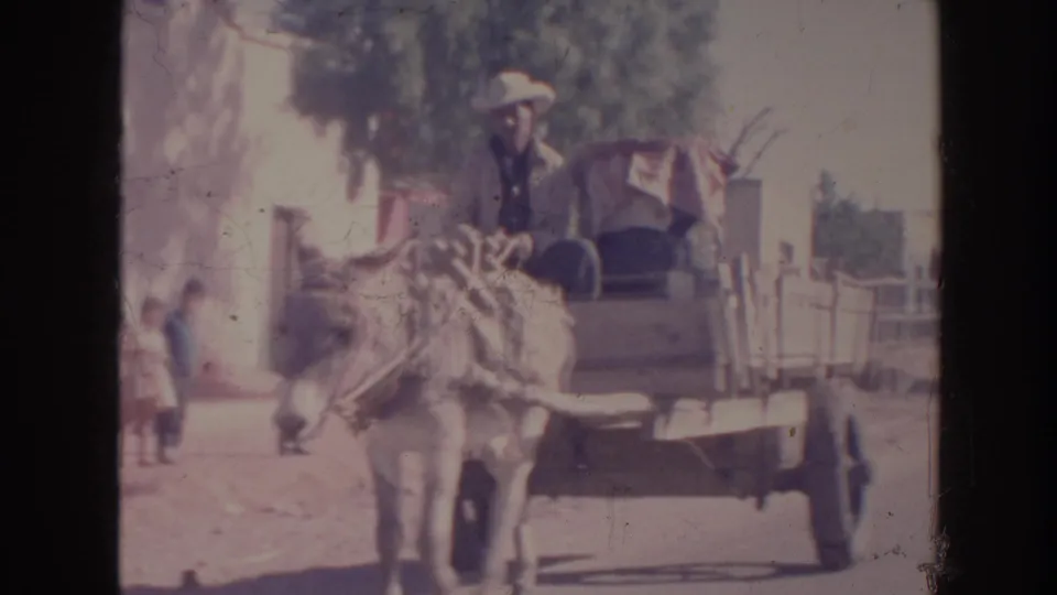 1961 8mm frame showing the donkey-drawn cart farther along the road, with the driver seated and buildings in the background.