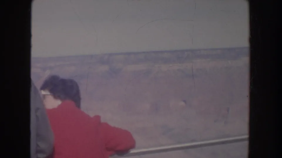 1962 Arizona home-movie frame showing a woman in a red jacket and white sunglasses near a railing above a canyon view.