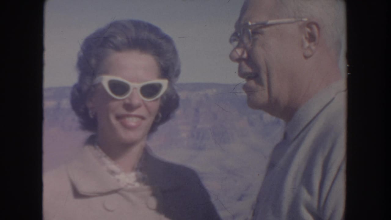 Smiling woman in white sunglasses stands beside an older man with a canyon behind them.