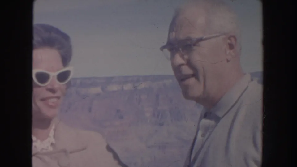 An older man in glasses and a light shirt faces the camera while the woman in sunglasses is partly visible at the canyon overlook.