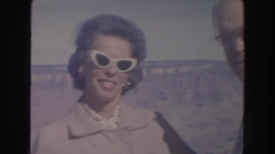 The woman smiles in white cat-eye sunglasses and a light coat, with the canyon behind her and the man partly cropped at the side.