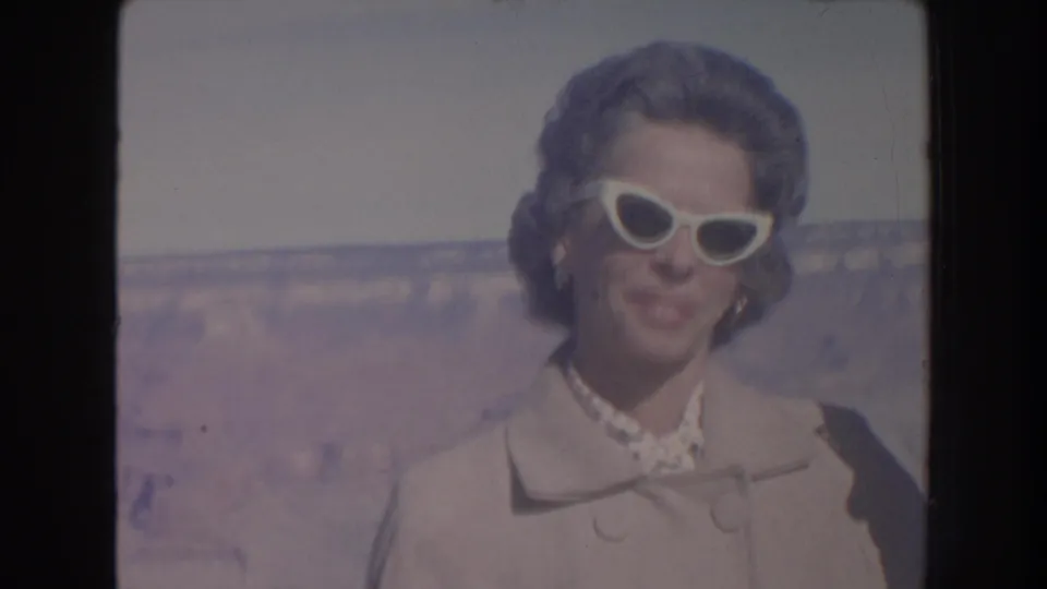 1962 Arizona home-movie frame showing a woman in a light coat, pearl necklace, and white cat-eye sunglasses with the Grand Canyon behind her.
