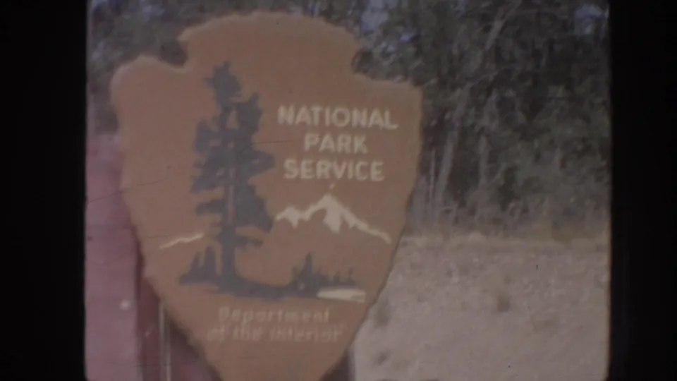 1962 Arizona 8mm frame showing a close view of a National Park Service arrowhead sign with trees and roadside ground behind it.