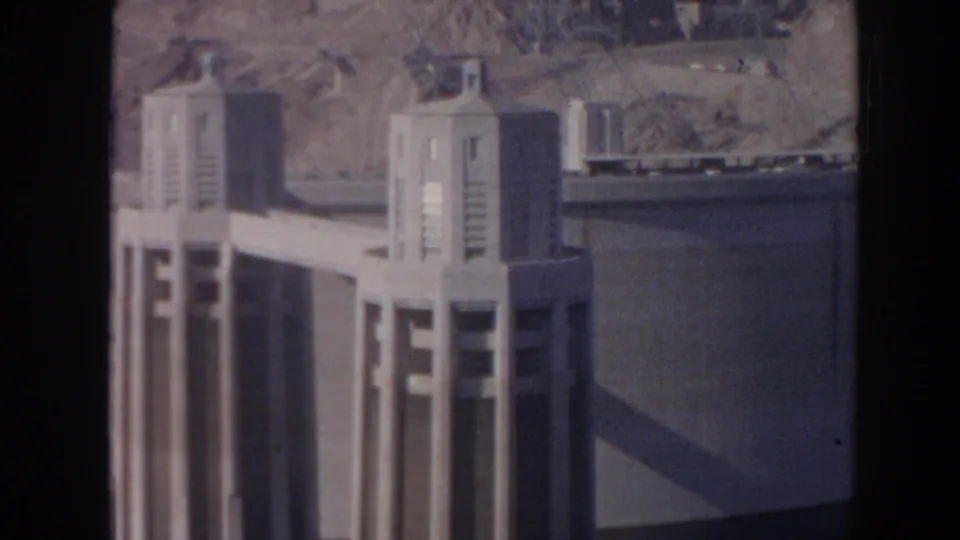 1962 8mm view of Hoover Dam showing a concrete intake tower, curved dam wall, dark water, and desert slope beyond.