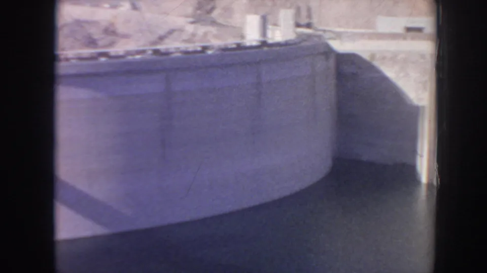 1962 8mm view of Hoover Dam's curved concrete wall, reservoir water, desert rock, and bright sunlit spillway area.