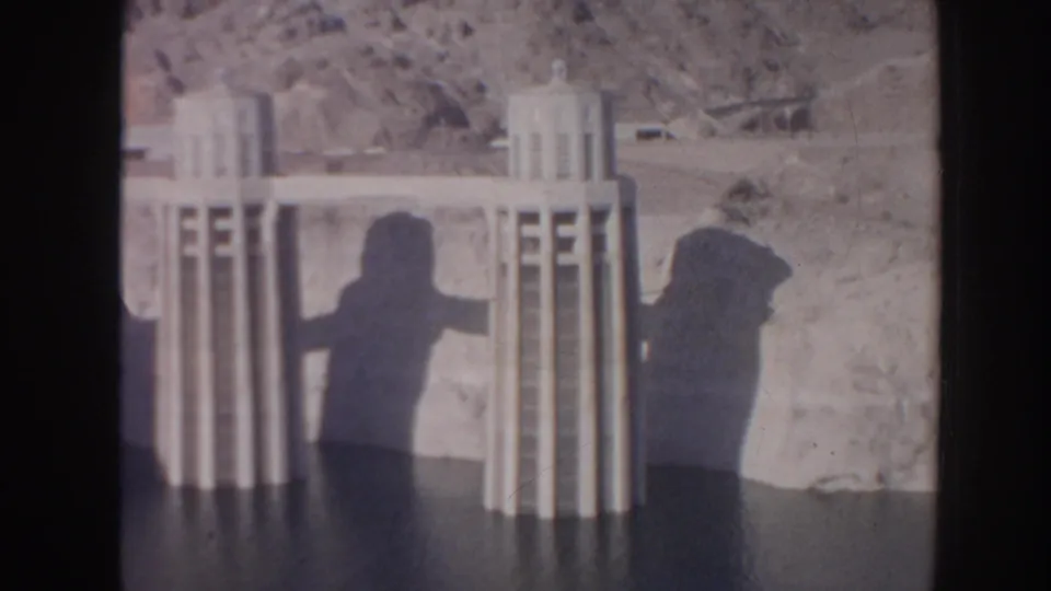 1962 Arizona home-movie frame of Art Deco-style dam towers, reservoir edge, rugged hillside, and visitor shadows cast on the structure.