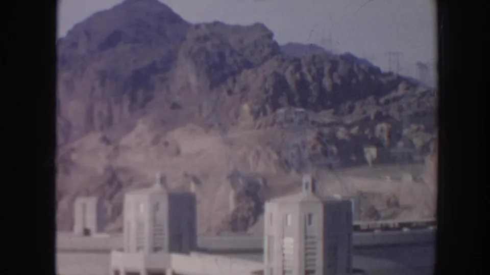 1962 Arizona frame showing concrete dam towers below rugged desert mountains, with soft color and visible 8mm film wear.