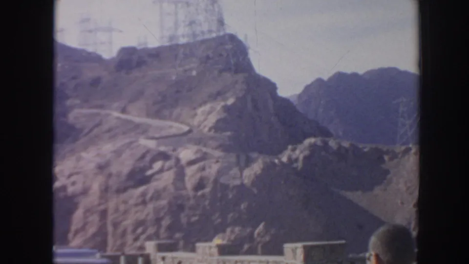 Hoover Dam area landscape with power lines, rocky slopes, and a partially visible visitor in the foreground.