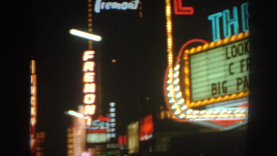 1960s home-movie frame showing Fremont neon near a theater marquee with glowing bulbs and partial show text.