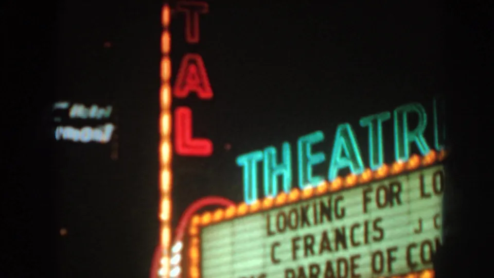 Soft 8mm nighttime view of a theater marquee advertising Looking for Love and Big Parade of Comedy, with nearby neon signs.
