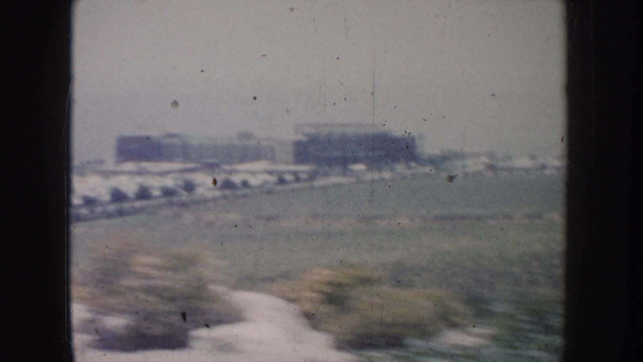 1962 California 8mm frame showing an open roadside field with distant low buildings and dark car-window edges.