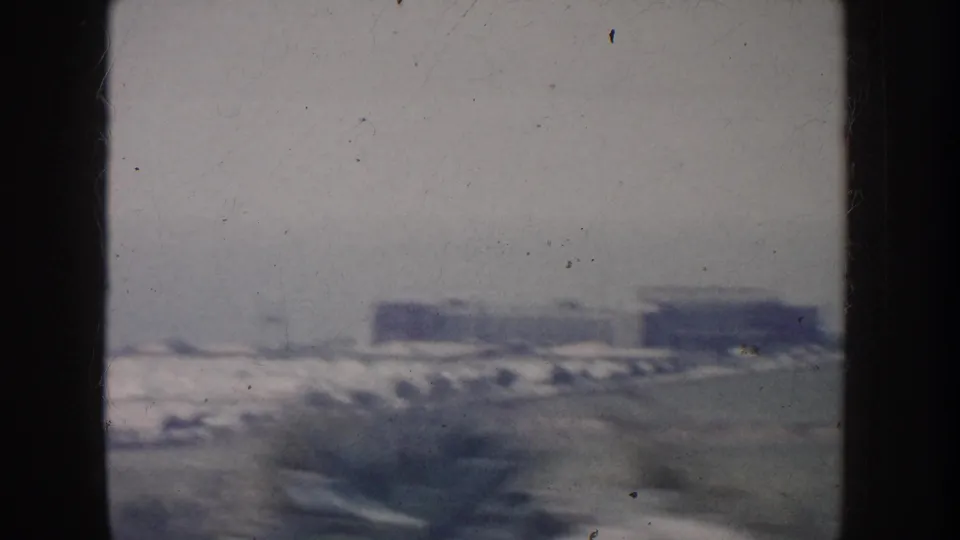 1962 California roadside view from a moving car, with open ground, distant buildings, and small cars along the far edge.