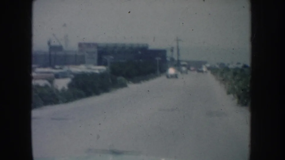 1962 California 8mm road scene with low buildings to the left, cars along the roadside, and vegetation at the curb.