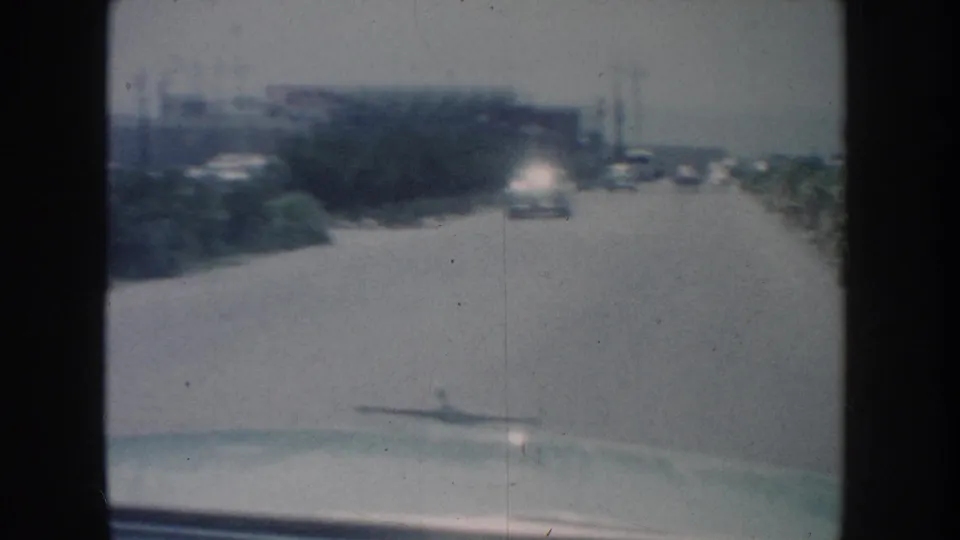 1962 California moving-car view over a pale hood, looking down a broad road with vehicles and low roadside buildings.