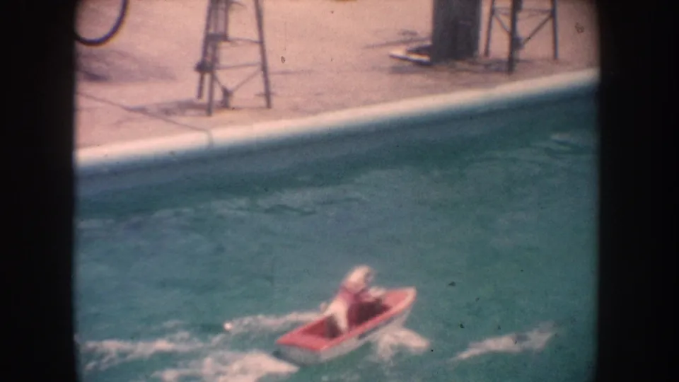 A dog in a red model speedboat moves across a blue-green swimming pool in California home-movie footage from 1962.
