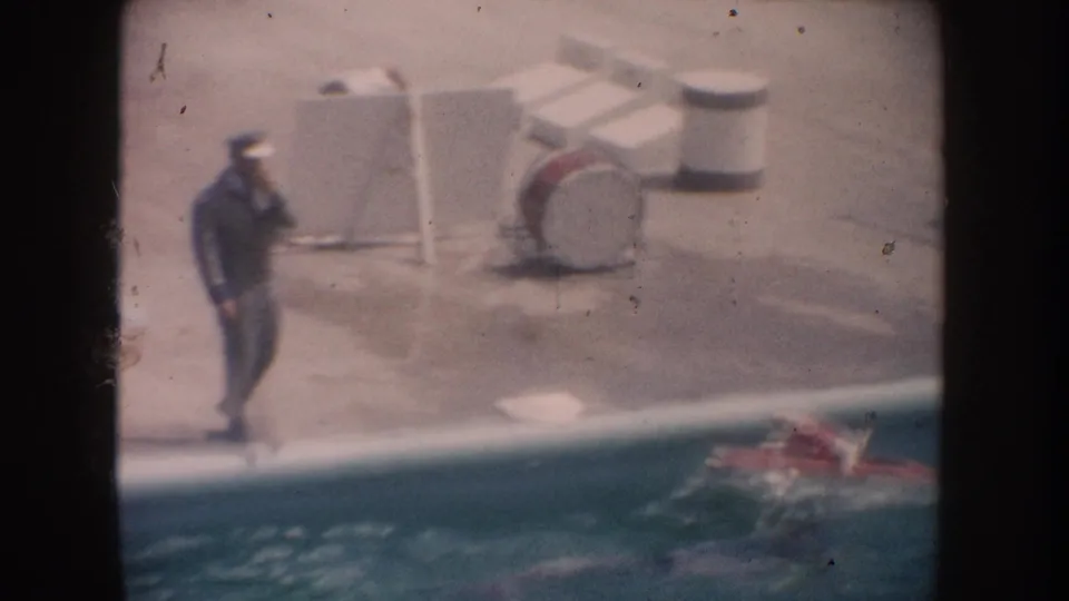 An adult stands near patio furniture on the pool deck while the dog in the red model boat moves along the water below.