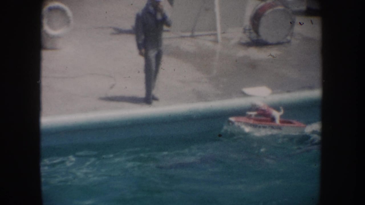 1962 California 8mm frame showing a poolside man and a small red motorized boat carrying a dog across the pool.