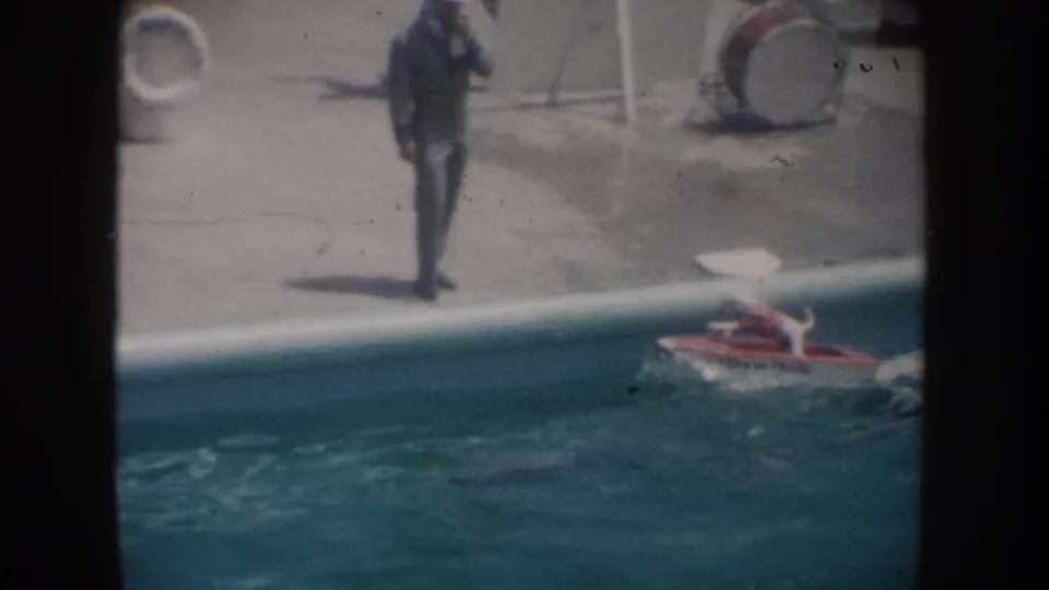 1962 California 8mm frame showing a poolside man and a small red motorized boat carrying a dog across the pool.