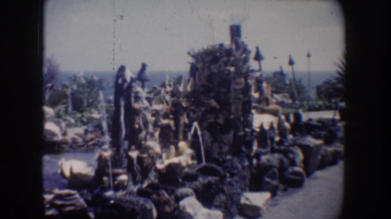 1962 California 8mm frame of a decorative rock grotto with fountains, clustered plants, and a coastal horizon in the background.