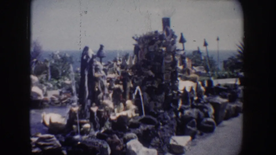 1962 California 8mm frame of a decorative rock grotto with fountains, clustered plants, and a coastal horizon in the background.
