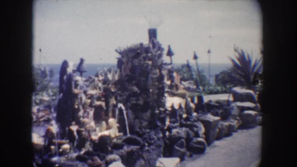 1962 California home-movie frame showing a rock fountain garden with water features, palm-like plants, and ocean or horizon beyond.
