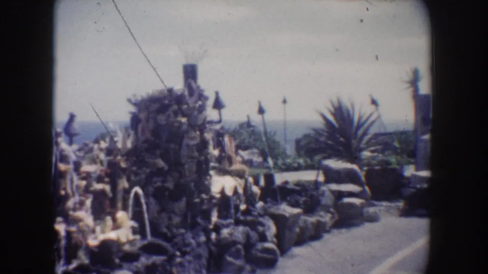 1962 California 8mm frame of a rock grotto fountain display beside a paved walkway, with plants and distant water visible behind.