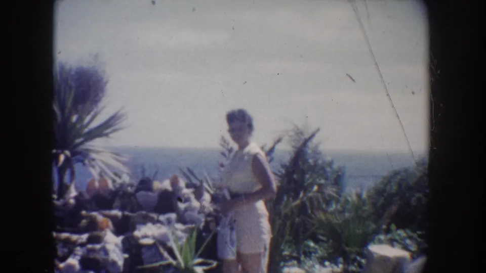 1962 California home-movie frame with a woman in a light dress walking through a coastal garden, with palms, rocks, plants, and ocean behind her.
