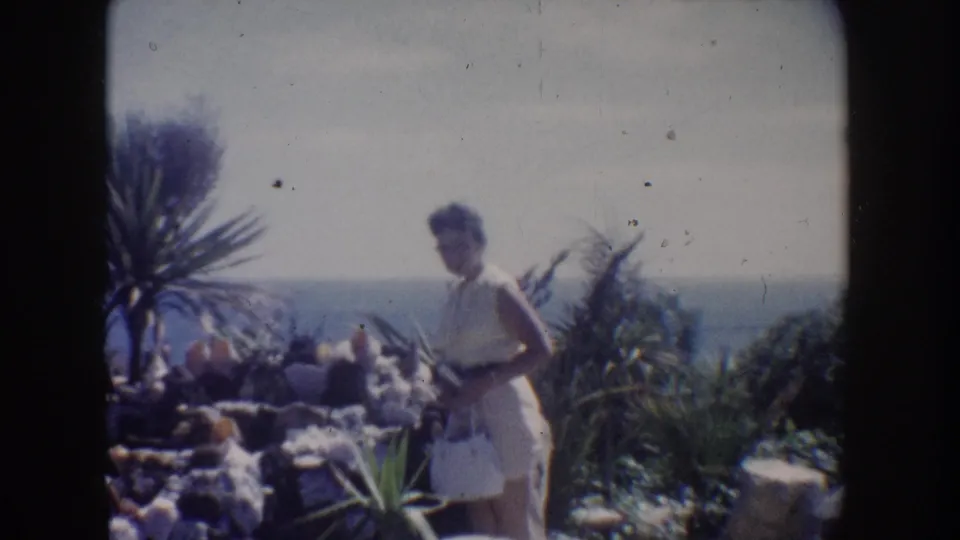 Vintage 8mm frame from California showing a woman in a light dress beside rocks and spiky garden plants, with palms and the ocean horizon behind her.