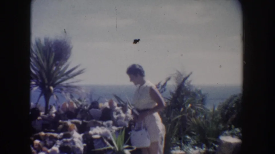 1962 California 8mm frame showing a woman in a pale dress beside rocky coastal planting with a yucca-like palm and ocean horizon.