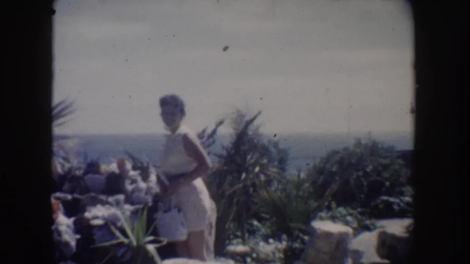 1962 California 8mm still with a woman holding a white handbag on a sunny coastal overlook, surrounded by rocks, plants, and sea haze.