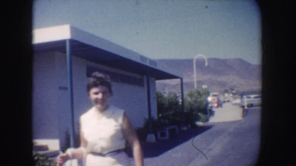 Adult woman in a white sleeveless dress walks beside a paved road near a mid-century flat-roofed building in California, 1962.