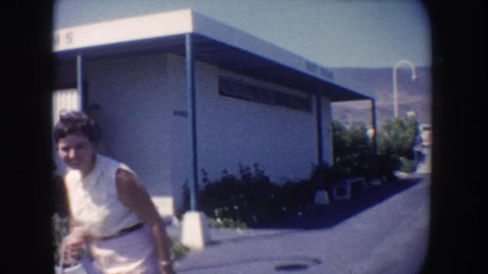 Smiling adult woman passes a mid-century flat-roofed building along a paved California roadside in 1962 home-movie footage.