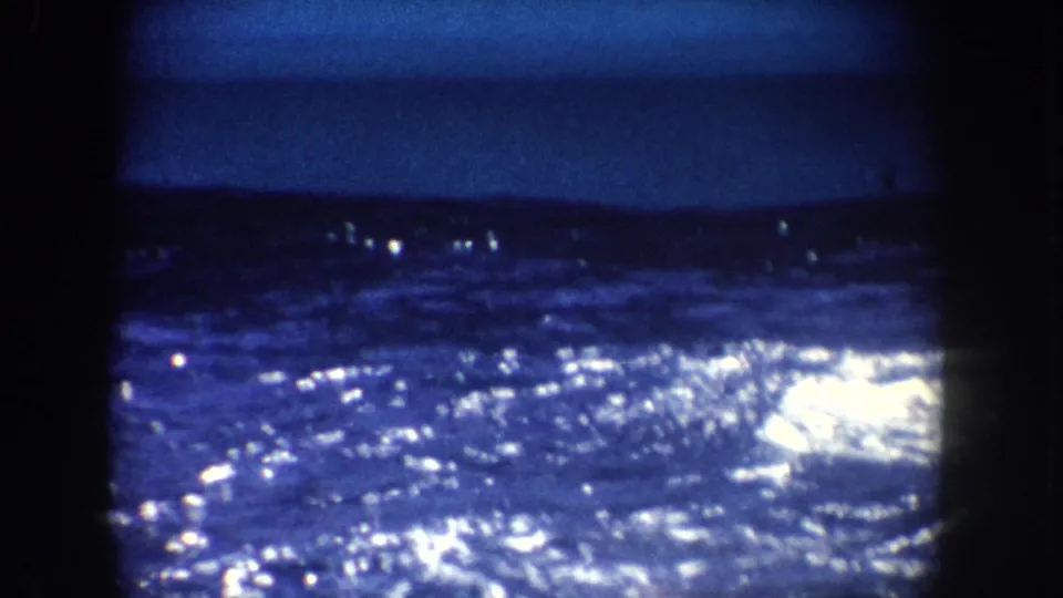 Blue-toned 8mm frame of rippling water and a distant dark shoreline or hills, with bright reflected highlights.