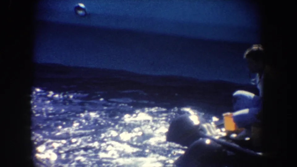 Blue-shifted 8mm frame showing sunlit water, distant hills, and partially visible people near the lower and right edges.