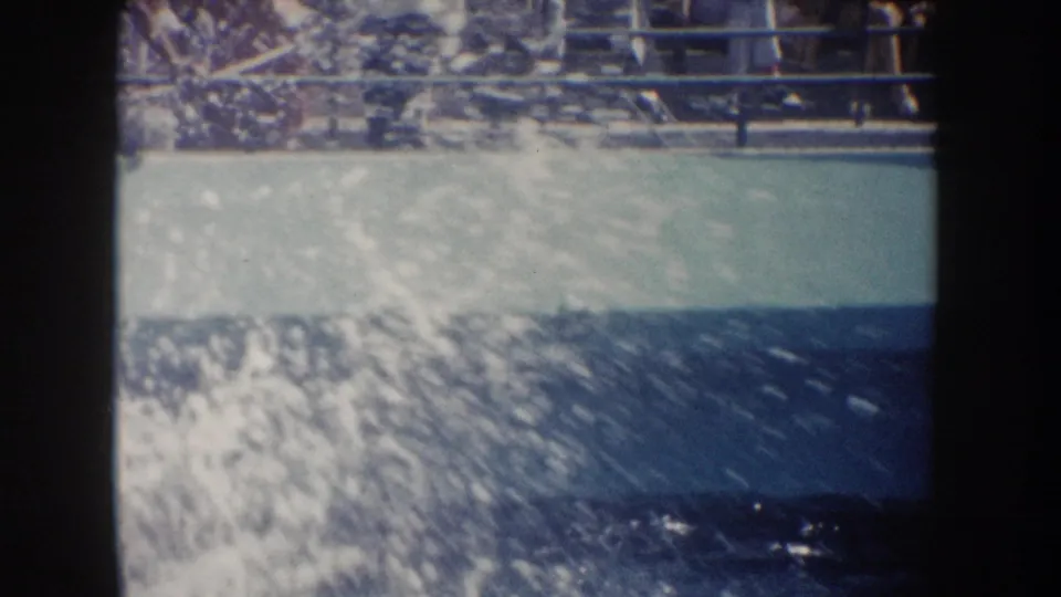 1962 California home-movie frame showing pool water, a broad splash, and spectators behind railings.