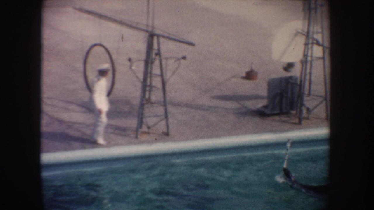 1962 California home-movie frame showing a white-uniformed trainer at poolside with a hoop, poles, and a seal partly visible in the water.